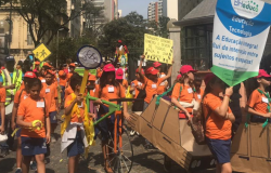 Alunos estão na rua vestidos de bermudas azul-marinho e camisetas cor de laranja, seguram cartazes, brinquedos e um estandarte, participando de um cortejo da educação.