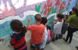 Cerca de 8 crianças e uma professora em uma escadaria. As crianças estão segurando pincéis e pintam um muro onde pode-se ver algumas flores vermelhas desenhadas