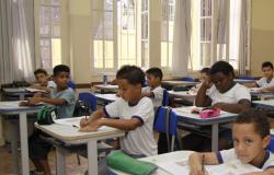 Cerca de 8 alunos estão em uma sala de aula com uniformes da escola, camisetas brancas.