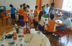 Cerca de 12 alunos vestidos com camisetas cor de laranja estão em uma sala com dois monitores fazendo trabalhos manuais com papelão, garrafas plásticas, canudos plásticos e outros.