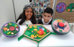 Uma menina e um menino sorriem para a foto, onde também se pode ver trabalhos feitos para a mostra científica, dois redondos e um em formato de pentágono.