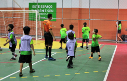 Futsal praticado crianças dentro de uma quadra poliesportiva. Presença de árbitro e de jogadores. 