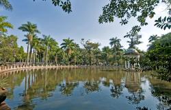 uma vista de uma lado da lagoa que fica no parque municipal Américo Renné Giannetti. Ela está rodeada por árvores e no meio dela tem um coreto. A foto foi tirada de dia, o céu está muio azul e sem nenhuma nuvem