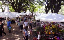 Foto de feiras com flores diversas em um ambiente muito arborizado, com várias pessoas passeando e realizando suas compras.