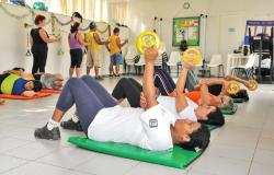 Homens e mulheres adultos se exercitam em sala da Academia da Cidade deitados em colchões de ginástica e levantando pesos e em pé puxando elásticos. Foto: Arquivo PBH