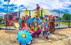 Doze membros de uma trupe de circo, com instrumentos musicais, malabares e outros acessórios, sorriem em frente à tenda de circo e à grade durante o dia. Foto: Divulgação