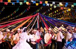 Sob um céu decorado por bandeirolas, mais de 15 integrantes de uma quadrilha junina dançam ao redor de um poste que com fitas coloridas, cada integrante segunrando uma delas. 