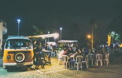 Kombi amarela é espaço de oferta de alimentos na praça Dino Barbieri; mais de dez pessoas frequantam o local, sentadas em cadeiras e conversando ou em pé. Ao fundo, outras barracas de alimentos. 