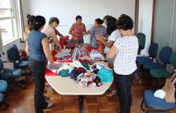 Cerca de sete mulheres em torno de uma mesa com várias roupas realizam oficina de convivência; 