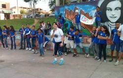 Crianças uniformizadas com blusas azuis da escola, e uma mulher falando no microfone posicionada na frente dessas crianças. 