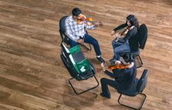 Três violinistas tocam, sentados em cadeiras. São quatro cadeiras e em uma delas 