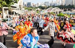 Cortejo de festa junina, com várias pessoas vestidas de roupas coloridas dançando quadrilha