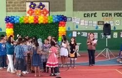 Mais de dez alunos con trajes de festa junina se organizem em frente a palco para se apresentarem. Ao lado, professora e ao fundo, os Dizeres "Xafé com poesia". 
