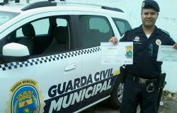 Guarda Municipal, ao lado de uma viatura, segura diplomas de cursos de capacitação em línguas