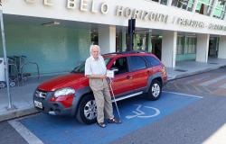 Millo da Conceição Nicolai, de 84 anos, tem uma deficiência na perna. Na foto, está encostado em seu carro, da cor vermelha, em uma vaga de estacionamento dedicada a pessoas com deficiência. 