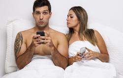 Foto promocional da peça com os dois atores utilizando o celular deitados em uma cama; mulher espia o celular do homem. Foto: Nello Aun