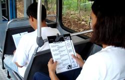 Dois passageiros de ônibus, sentados, lêem textos do Projeto Leitura para Todos, afixados na cadeira em frente ao passageiro. 