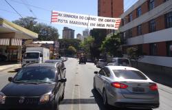 Carros transitam na Avenida Uruguai, próximo ao cruzamento com Avenida Nossa Senhora do Carmo; local tem faixa com os dizeres: Av. Nossa Senhora do Carmo em obras. Pista marginal em meia pista.