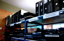 Vários monitores e outras partes de computadores e eletrônicos em prateleira de Centro de Recondicionamento da Prodabel. Em cada setor, uma polaca indicativa. A placa legível diz: recondicionamento de computadores.