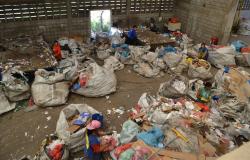 Galpão de reciclagem de lixo, com muitos sacos de lixo e uma mulher cercada de copos plásticos.