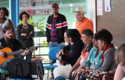 Dois homens com tocam violôes e sete pessoas, das quais quatro estão em pé, assistem ou cantam junto, atividade do projeto Varanda Musical, do Centro Cultural Alto Vera Cruz.