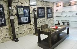 Sala do Museu da Inconfidência com mesa histórica e fotos e documentos expostos em parede de pedra e estante de vidro. 