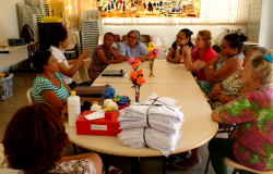 Nove mulheres reunidas em torno de mesa com panos, linhas e cadernos.