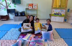 Professora e duas crianças sentados em espaço literário de escola e seguram almofadas escritas