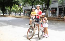 Homem de bicileta e capacete com filho no colo de capacete e garota ao lado, com bicileta e capacete.