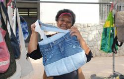 Mulher mostra bolsa artesanal feita com tecido e partes de calça jeans na banca onde vende produtos semelhantes.