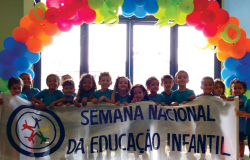 Cerca de 16 crianças seguram a faixa com os dizeres: "Semana nacional da educação infantil" tendo atrás uma guirlanda de balões. 