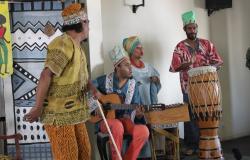 Três homens e uma mulher trajados com roupa africana em um palco. Um toca um violão, outro toca um tambor e a mulher toca um xilofone. O outro homem dança.