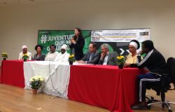 Nove pessoas compondo a mesa de abertura da Conferência de Igualdade Racial; com destaque para a secretária municipal de Políticas Sociais Maíra Colares de pé, ao centro.