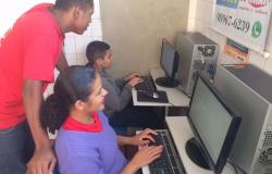 Monitor atuando em telecentro, auxiliando dois alunos que estão em frente ao computador