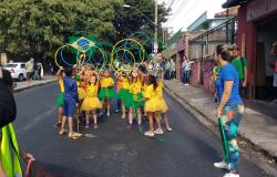 Mais de dez meninas de camiseta amarela e saias verdes ou amarelas e um menino de bermuda e casaco azul fazem movimentos com bambolês verdes, amarelos ou azuis em passeata na rua. Junto ao grupo, ainda havia a bandeira do Brasil.