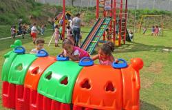 Cerca de quinze crianças, acompanhadas por educadores infantis, em pátio de Umei; uma brincam em túnel colorido, outras em escorregador e outras em cadeiras de balanço.