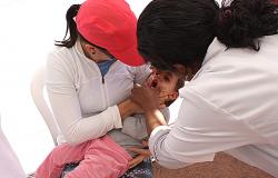 Filho no colo de mãe recebe vacina oral de agente de saúde.