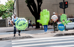 Campanha de trânsito é realizada em faixa de pedestre. Dois personagens: um mímico segurando um guarda chuvas escrito "Obrigado por me respeitar" e uma mão verde, que representa o "pare" do semáforo. Além de um guarda municipal de costas.