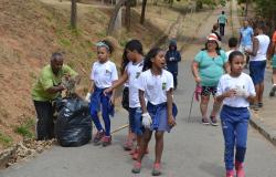 Quatro alunos de escola municipal caminham no Parque Vila Pinho durante o dia; ao lado, funcionário recolhe lixo; atrás, cidadãos também fazem caminhada. 