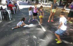 Cinco crianças desenham no chão de rua interditada; aduldos observam. 