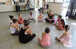 Nove garotas fazem aula de balé, sentadas em círculo, orientadas por professora; ao fundo da sala, uma garota está sentada no colo de responsável. 