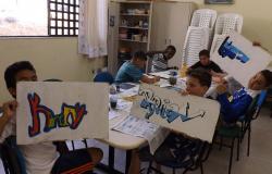 Cinco alunos, sentados em mesa, fazem oficina de grafite; três deles mostram seus desenhos coloridos. 