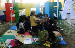 Cerca de nove crianças manuseiam livros; atrás delas está escrito "Leitura" com letras coloridas e gigantes. 