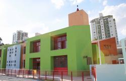 A foto mostra uma construção, paredes pintadas de verde claro e azul claro, grades de ferro pintadas de vermelho, duas janelas grandes e 2 portas de vidro. Atrás da construção vê-se dois prédios bem altos.