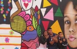 Três alunos posam à frente de muro com grafite. 