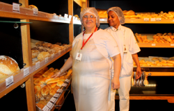 Duas mulheres com roupa branca, avental e touca, à frente de estante de padaria, com diversos pães.