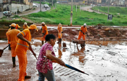 Sete funcionarios da SLU realizam limpeza de rua após chuva e uma senhora passa pelo local.