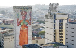 Mulher afro com turbante na cabeça grafitada em lateral de edifício. Em sua cintura, os dizer "coragem". Ao fundo, mais prédios e cenário urbano.