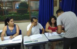 Três pessoas sentadas em mesas escolares; à frente, adulto preenche formulário, de pé, uma etapa da eleição para gestores escolares.