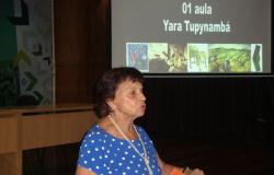 Palestrante Yara Tupinambá em pé e, logo atrás, uma imagem projetada de sua palestra.